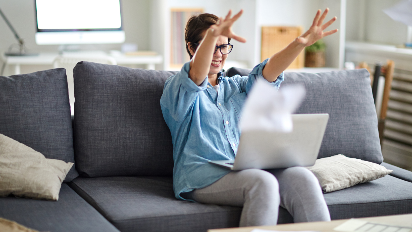 A frustrated woman with bipolar disorder experiencing irritability, sitting on a couch with her laptop and throwing papers in the air in frustration, conveying emotional distress and difficulty managing mood.