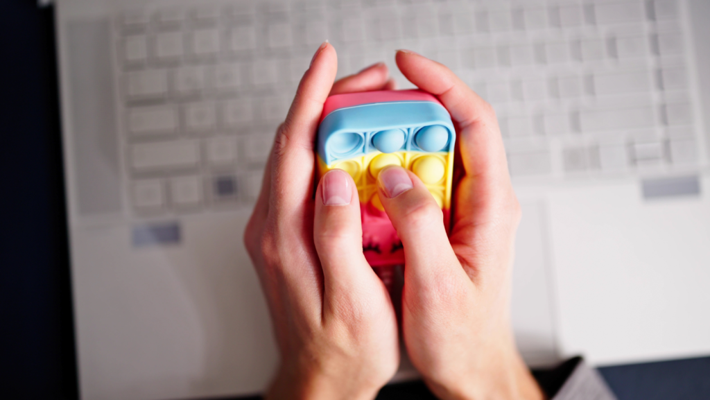 Close-up of a person's hands holding and pressing a colorful sensory fidget toy while sitting at a desk, using it to manage anger and irritability.