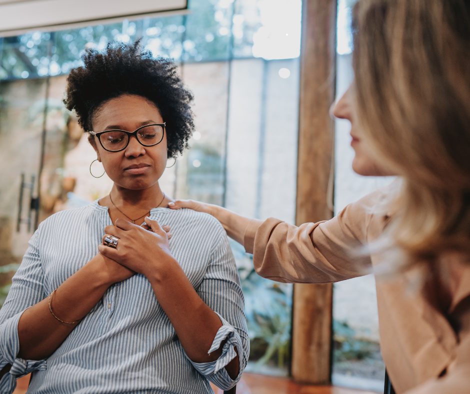 A woman sits with her hand on her chest, visibly processing her emotions with a thoughtful expression. Another person gently places a supportive hand on her shoulder, symbolizing empathy and the importance of seeking help. This image reflects emotional connection and the journey to regulate your emotions in a supportive environment.