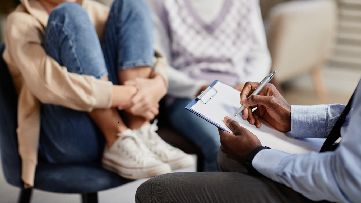 A mental health professional taking notes during a session with a teenage patient, who is sitting with knees pulled up, looking distressed.