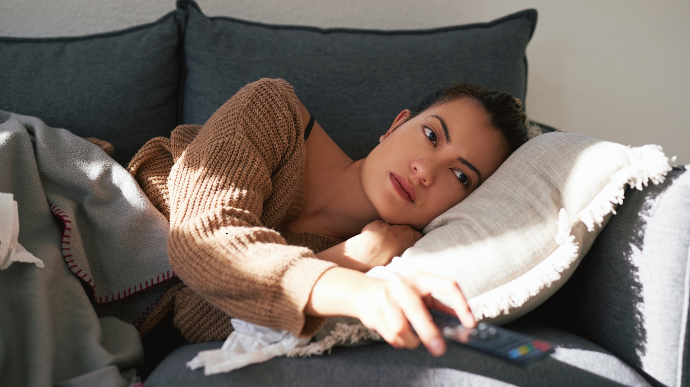 A young woman lying on a couch, holding a TV remote, with a contemplative expression, symbolizing the emotional weight of atypical depression.