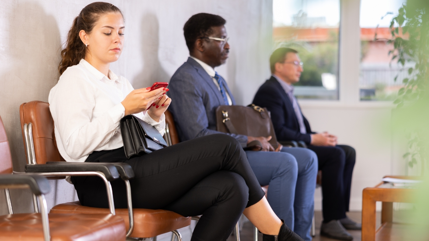 A woman sitting in a waiting room, looking at her phone, with others seated nearby, symbolizing the experience of preparing for your first psychiatric visit.