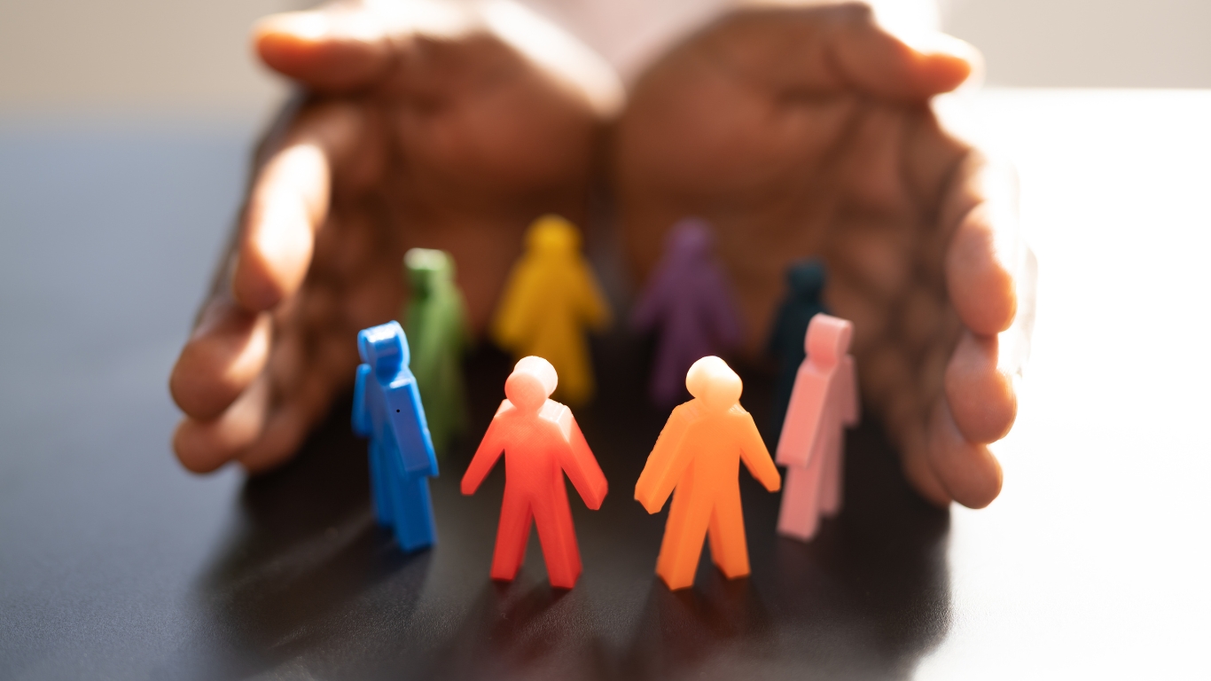 Close-up of diverse, multicolored human-shaped figures arranged in a circle with a pair of hands gently cupped around them, symbolizing protection, inclusion, and cultural competence in mental health care.
