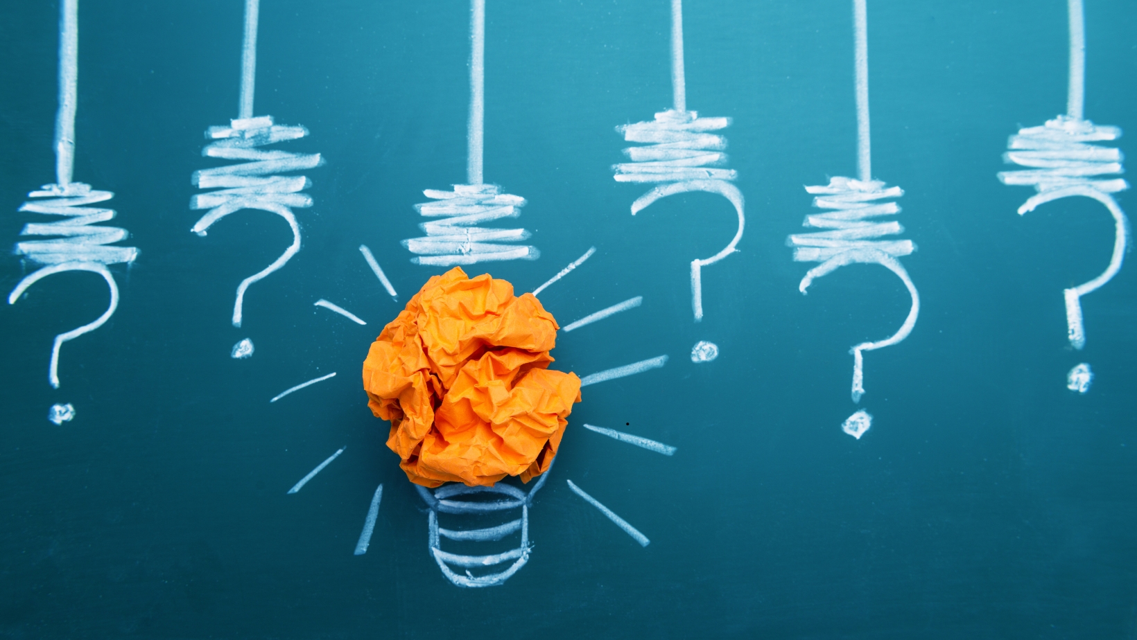 Concept image of catastrophic thinking, showing crumpled orange paper inside a chalk-drawn light bulb surrounded by question marks on a chalkboard.