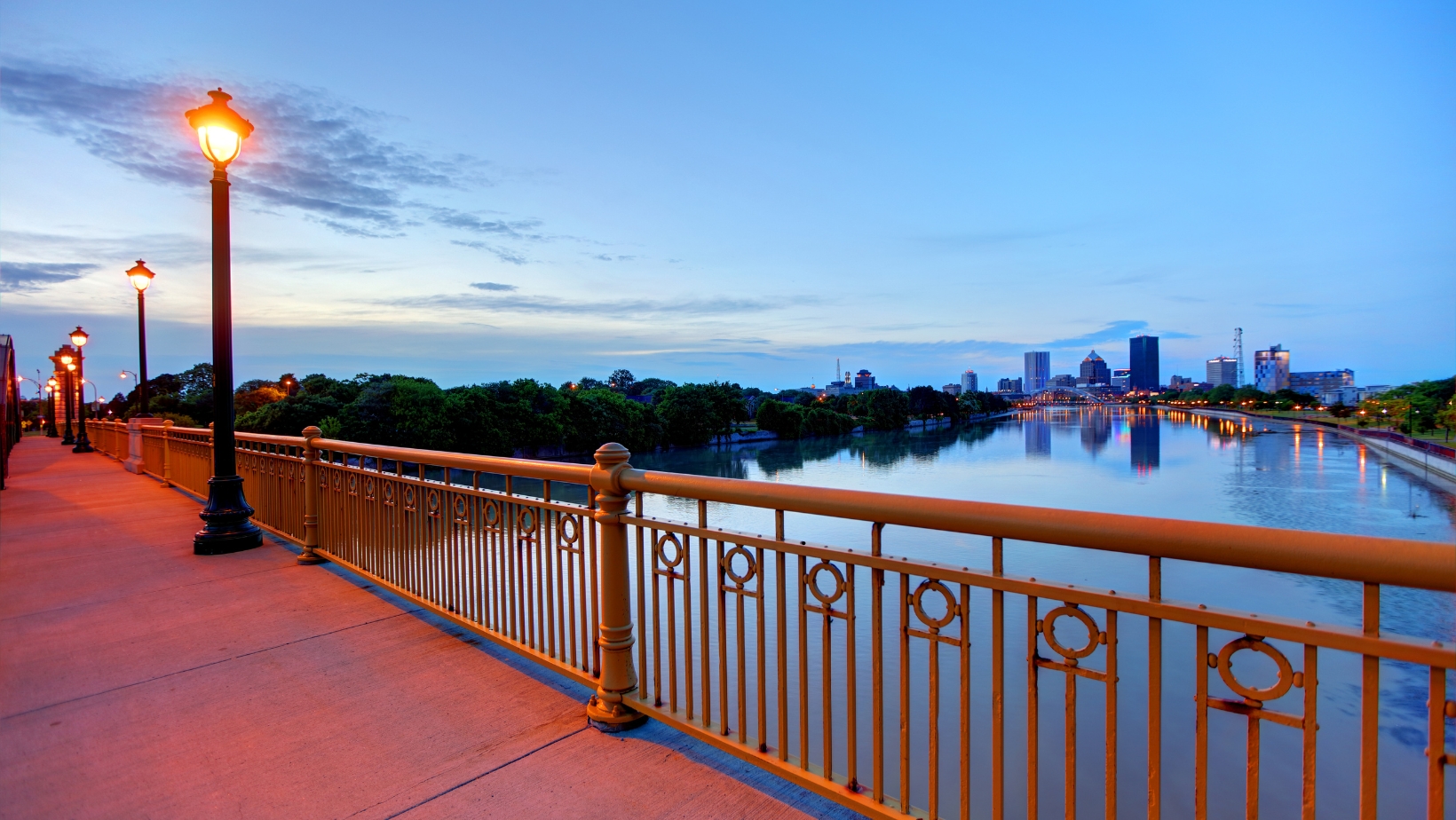 View of the Rochester, NY skyline at dusk with city lights reflecting on the Genesee River, seen from a pedestrian walkway lined with street lamps.