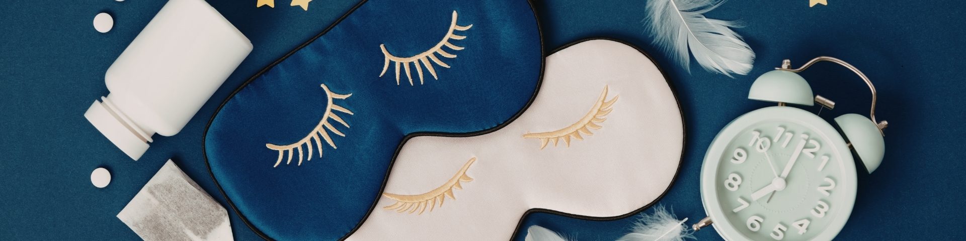 Flat lay of sleep items on a dark blue background, including two eyelash sleep masks, an alarm clock, earplugs, and a pill bottle, suggesting a bedtime routine and sleep hygiene.